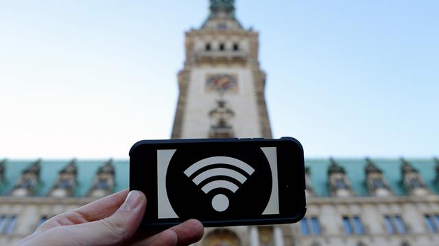 Eine Hand hält ein Schild mit einem WLAN-Symbol, im Hintergrund das Hamburger Rathaus.