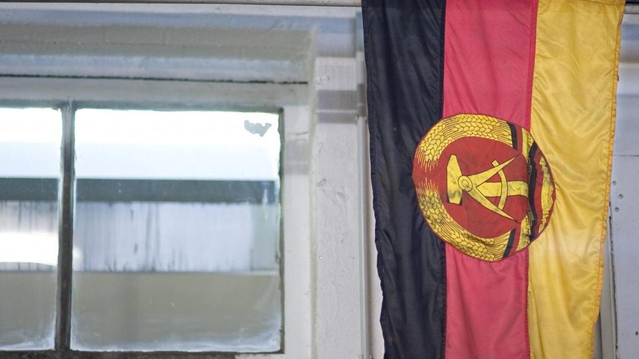 Eine DDR-Fahne mit den Symbolen Hammer und Zirkel hängt in einem Raum mit Fenster. Eine DDR-Fahne mit den Symbolen Hammer und Zirkel hängt in einem Raum mit Fenster.