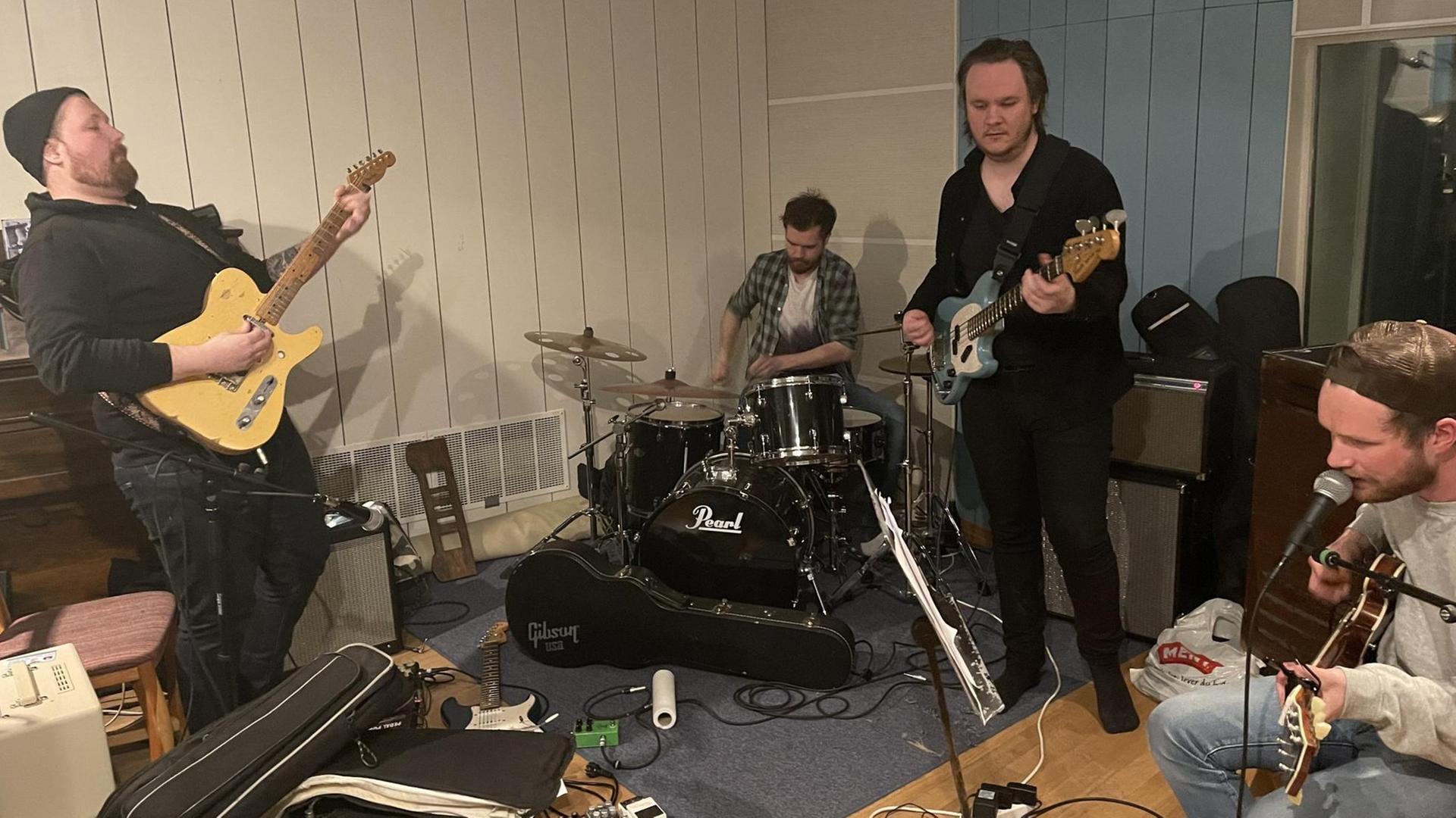Zwei Gitarristen, ein Bassist und ein Schlagzeuger spielen in ihrem Probenraum: Sie stehen bzw. sitzen zwischen Gitarrenverstärkern, Koffern, Kabeln. Zwei Gitarristen, ein Bassist und ein Schlagzeuger spielen in ihrem Probenraum: Sie stehen bzw. sitzen zwischen Gitarrenverstärkern, Koffern, Kabeln.