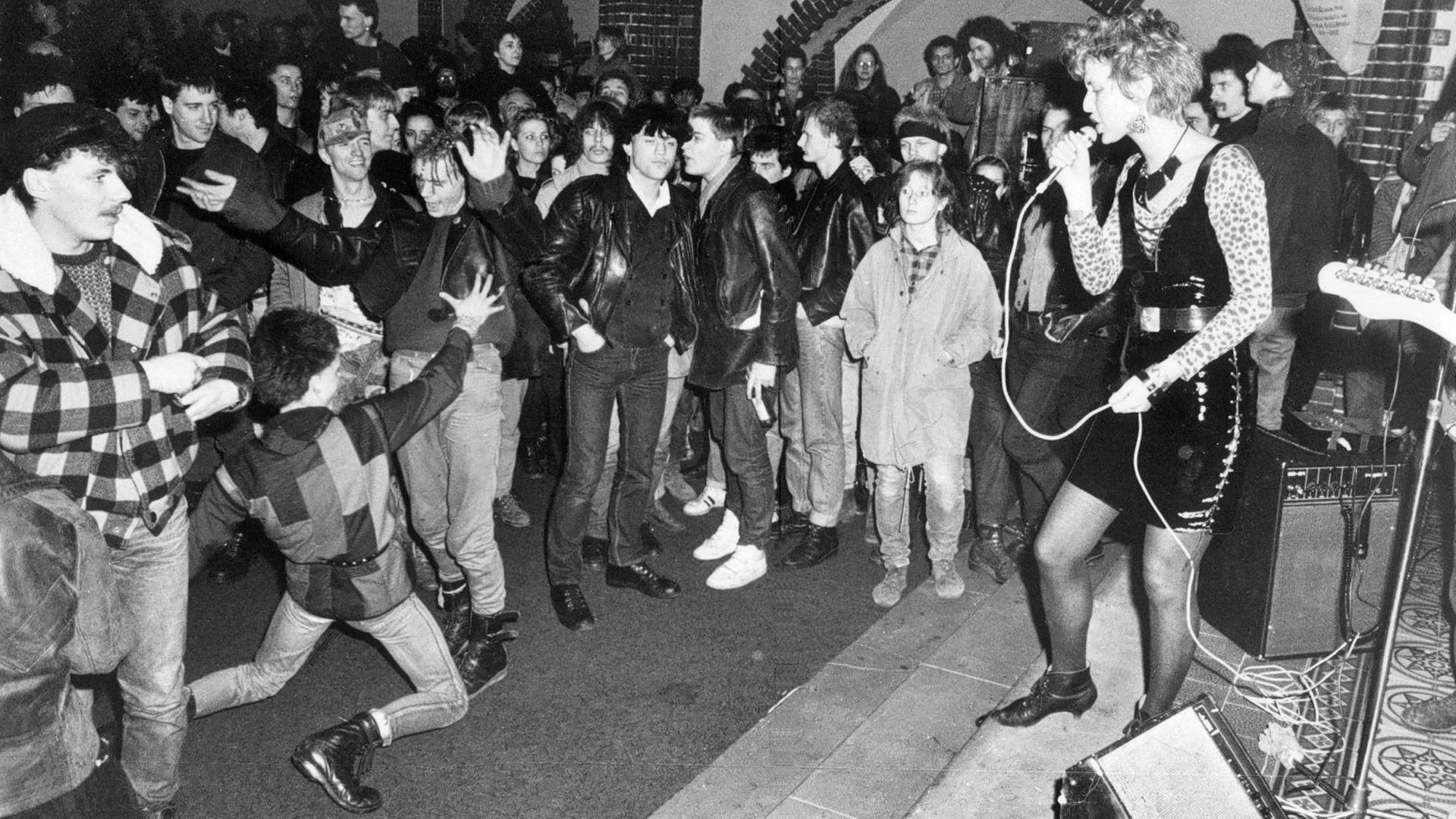 Auf der schwarzweiss Aufnahme ist das Benefizkonzert in der Berliner Erlöserkirche 1988 zu sehen. Eine Sängerin steht vor einer tanzenden Masse aus jungen Menschen.