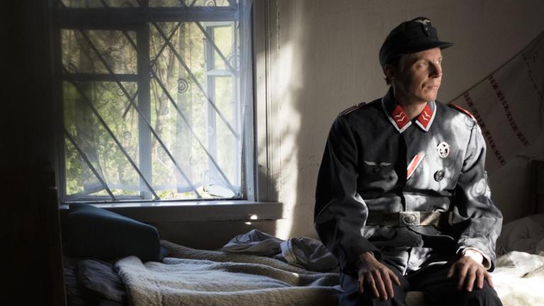 Sebastian Heinzel sitzt in Wehrmachtsuniform auf einem kargen Bett und blickt melancholisch ins Off.