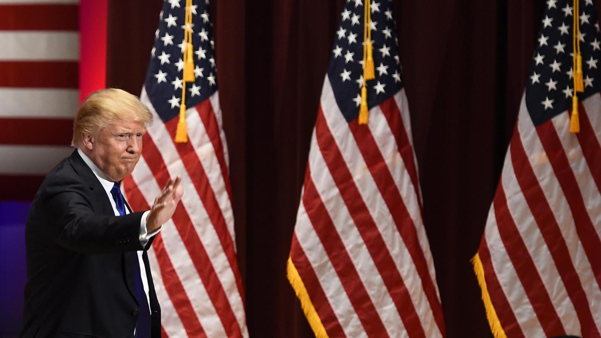 Man sieht Donald Trump, der winkt, im Hintergrund drei große US-Fahnen. Man sieht Donald Trump, der winkt, im Hintergrund drei große US-Fahnen.