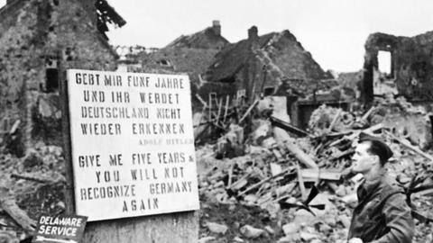 "Gebt mir fünf Jahre und ihr werdet Deutschland nicht wiedererkennen" steht in Deutsch und Englisch auf einem Schild vor einem Trümmerberg, ein Mann liest das Schild, schwarz-weiß-Aufnahme.