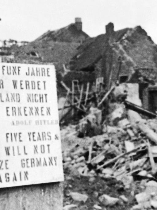 "Gebt mir fünf Jahre und ihr werdet Deutschland nicht wiedererkennen" steht in Deutsch und Englisch auf einem Schild vor einem Trümmerberg, ein Mann liest das Schild, schwarz-weiß-Aufnahme.