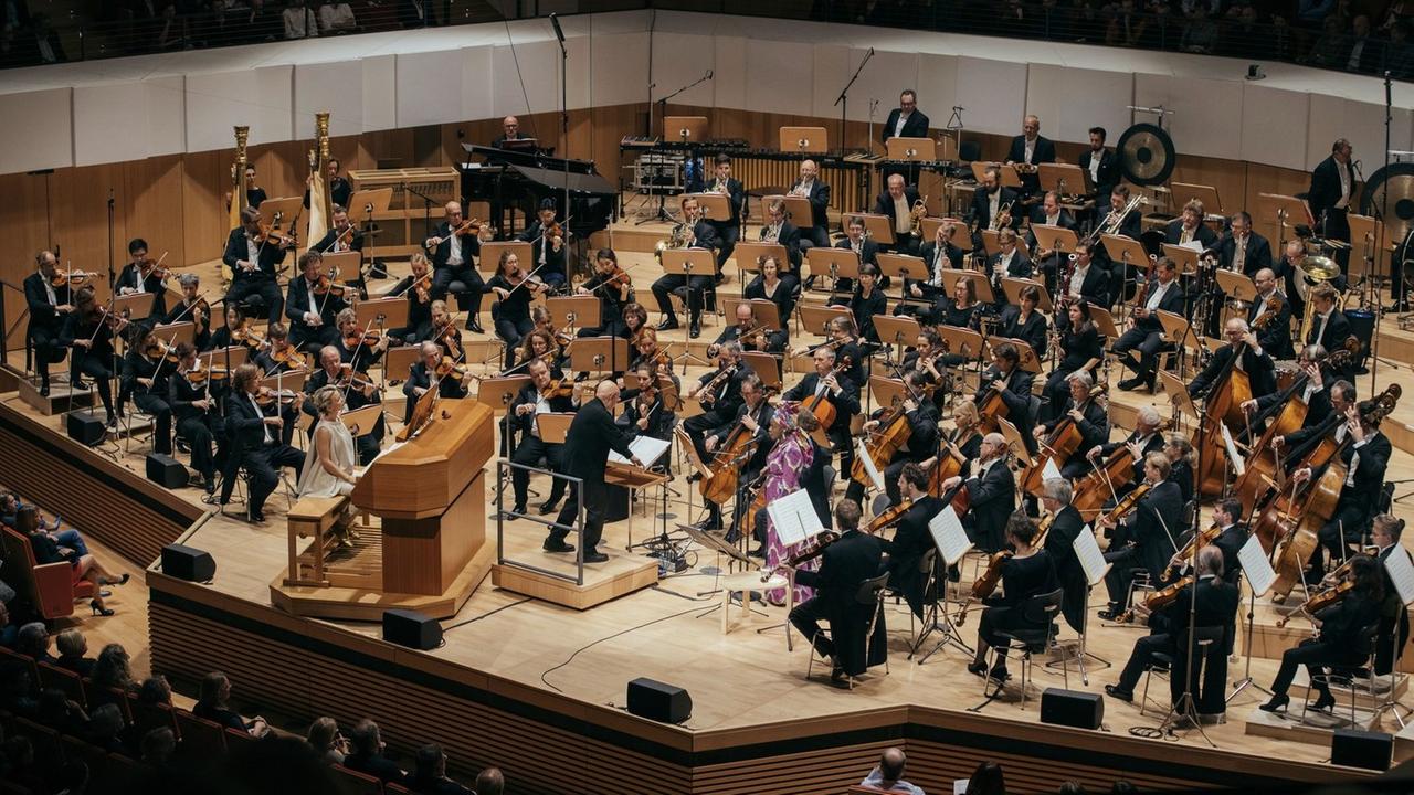 Über 100 Musikerinnen und Musiker sitzen in Anzug und Kostüm mit ihren Instrumenten auf einer Bühne, vorne in der Mitte steht der Dirigent, neben ihm sitzt eine Frau an einer Orgel.