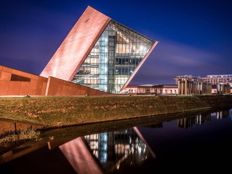 Der Museumsbau des Architekturbüro Kwadrat in Gdynia in Form eines schräg aus dem Boden ragenden und teilweise verglasten Würfels in nächtlicher Beleuchtung.