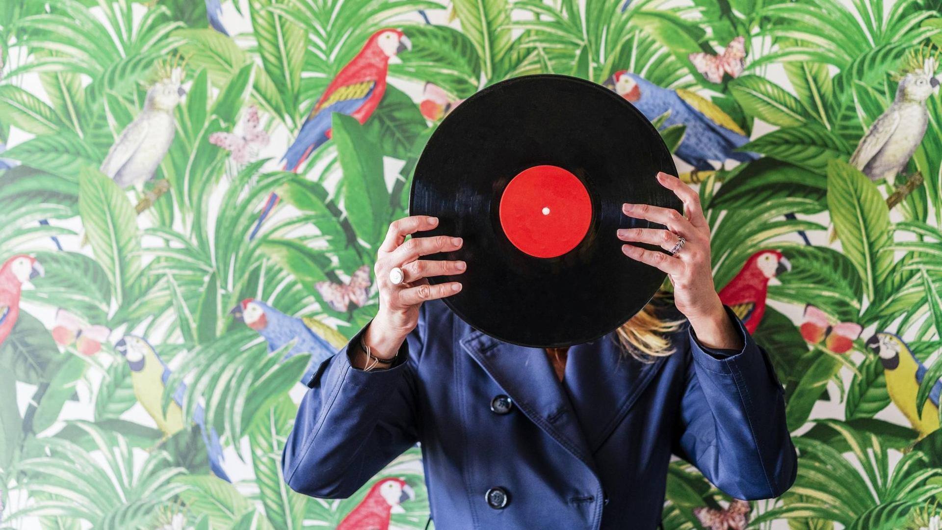 Eine Person vor einer Tapete mit Dschungeldekor bedeckt den Kopf mit einer Vinylschaltplatte. Eine Person vor einer Tapete mit Dschungeldekor bedeckt den Kopf mit einer Vinylschaltplatte.