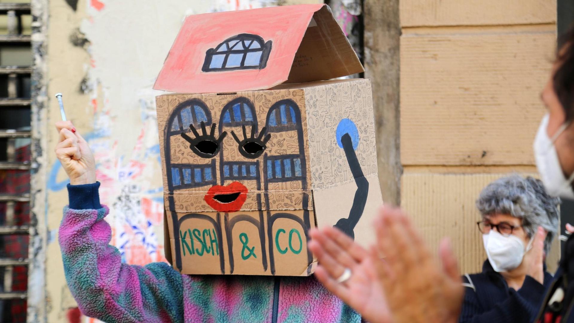 Ein Demonstrant hat sich einen zum Haus umgestalteten Pappkarton auf die Schultern und über seinen Kopf gesetzt. Der Pappkarton stellt das Haus dar, in dem sich die Räume von Kisch & Co. bislang befanden Ein Demonstrant hat sich einen zum Haus umgestalteten Pappkarton auf die Schultern und über seinen Kopf gesetzt. Der Pappkarton stellt das Haus dar, in dem sich die Räume von Kisch & Co. bislang befanden