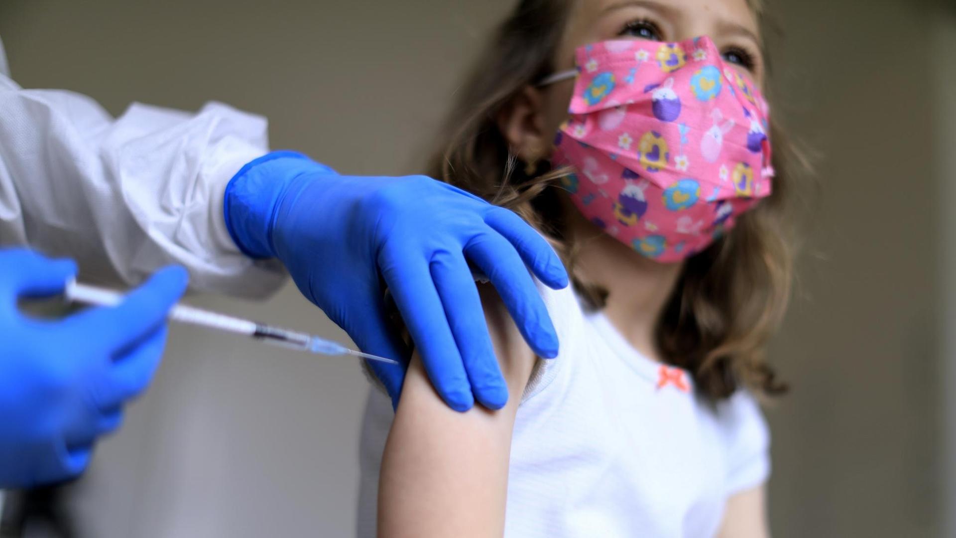 Ein Mädchen mit pinker Alltagsmaske bekommt eine Impfung.
