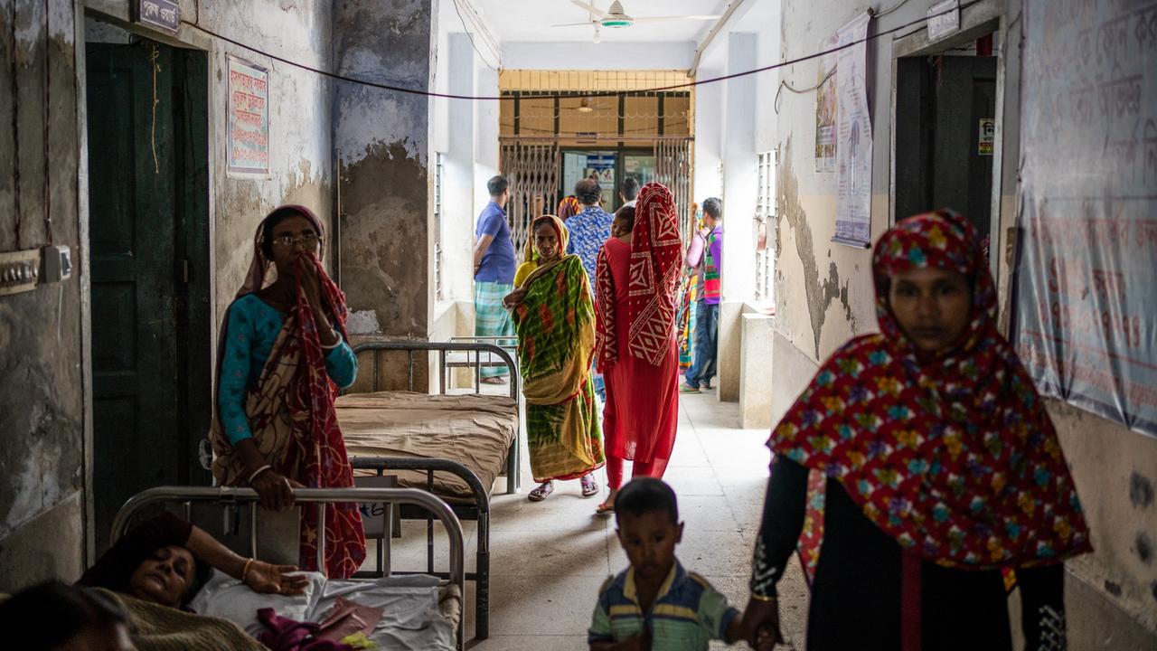 Ein Gang im Krankenhaus in Khulna, links liegt eine Patientin im Bett, rechts geht eine Mutter mit Kind an der Hand. Im Hintergrund wartende Angehörige Ein Gang im Krankenhaus in Khulna, links liegt eine Patientin im Bett, rechts geht eine Mutter mit Kind an der Hand. Im Hintergrund wartende Angehörige