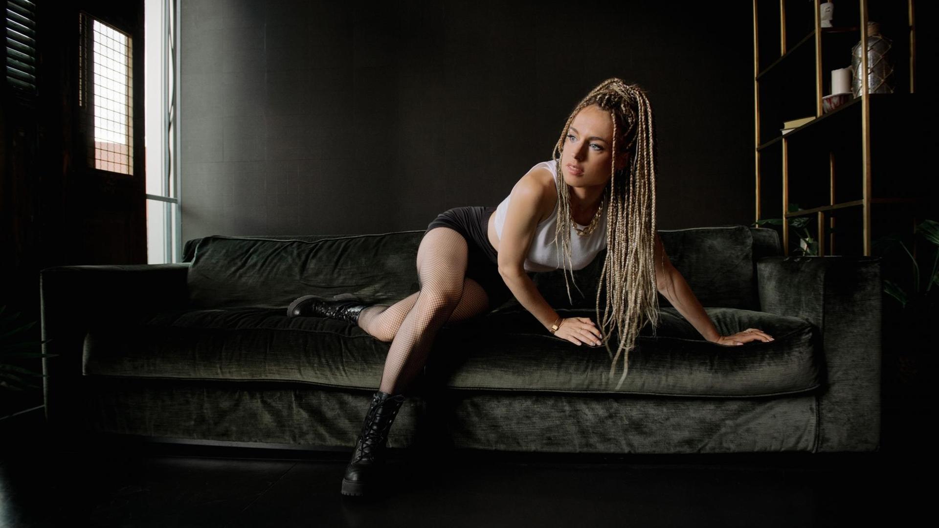 Marie Chain erhebt sich von ihrer schwarzen Couch, wobei ihre langen, schwarz-blonden Rastazöpfe über ihre Schultern fallen. Marie Chain erhebt sich von ihrer schwarzen Couch, wobei ihre langen, schwarz-blonden Rastazöpfe über ihre Schultern fallen.