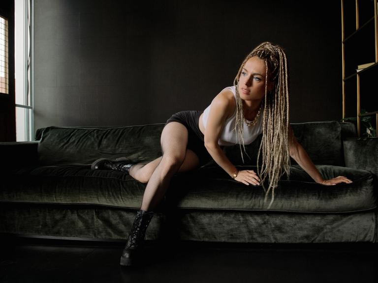 Marie Chain erhebt sich von ihrer schwarzen Couch, wobei ihre langen, schwarz-blonden Rastazöpfe über ihre Schultern fallen.