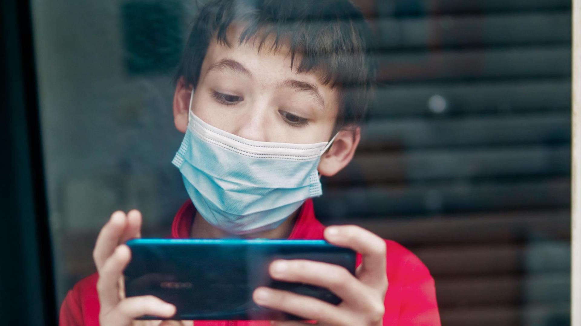 Ein Teenager mit medizinischer Maske benutzt hinter einem Fenster ein Smartphone. Ein Teenager mit medizinischer Maske benutzt hinter einem Fenster ein Smartphone.