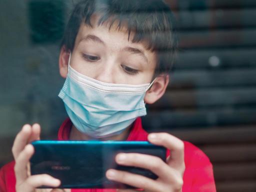 Ein Teenager mit medizinischer Maske benutzt hinter einem Fenster ein Smartphone.