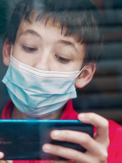 Ein Teenager mit medizinischer Maske benutzt hinter einem Fenster ein Smartphone.