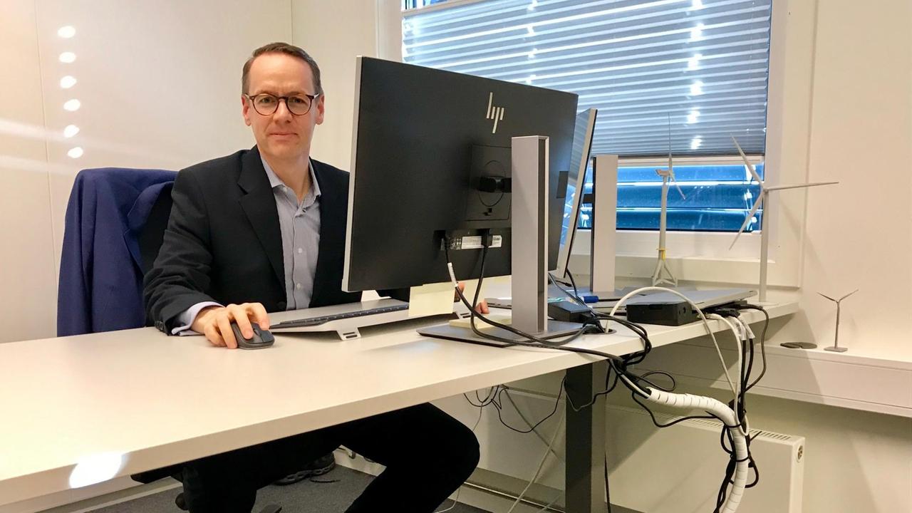 Ein Mann mit Brille sitzt im geöffneten Jacket an seinem Schreibtisch vor dem PC, die Maus in der Hand und schaut in die Kamera. Der Schreibtisch und der ganz in weiß gehaltene Raum sind ansonsten völlig leer. Ein Mann mit Brille sitzt im geöffneten Jacket an seinem Schreibtisch vor dem PC, die Maus in der Hand und schaut in die Kamera. Der Schreibtisch und der ganz in weiß gehaltene Raum sind ansonsten völlig leer.