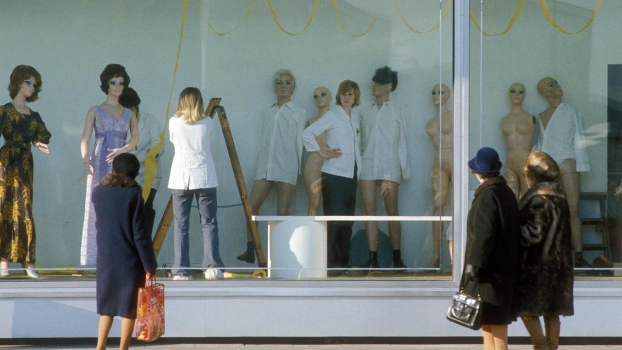 Eine Farbaufnahme zeigt Kunden am Schaufenster des Centrum Warenhaus auf dem Alexanderplatz in Ostberlin 1985. Hinter dem Schaufenster arrangiert und bestückt eine Mitarbeiterin die Modepuppen.