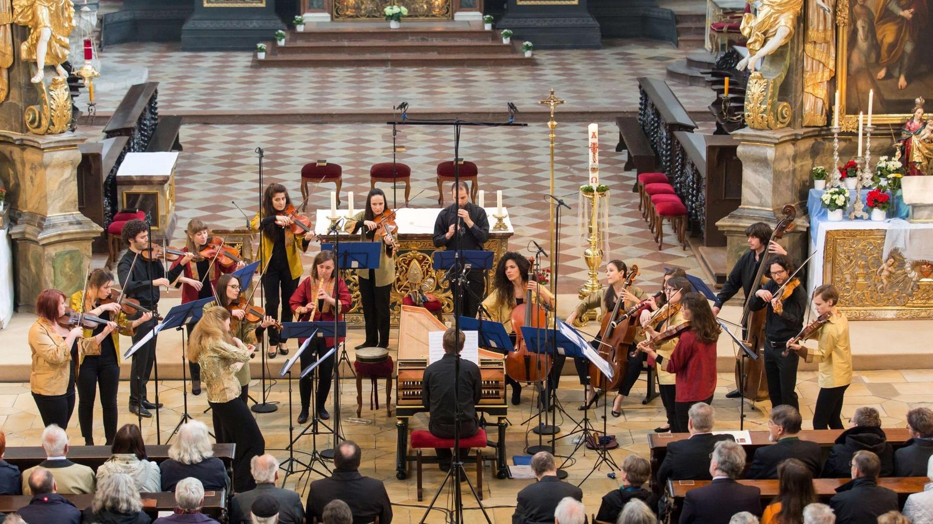 Die Musikerinnen und Musiker des Barockorchesters der Europäischen Union spielen überwiegend stehend im Altarvorraum einer Kirche. In deren Mitte sitzt der Cembalist an seinem Instrument. Der Boden ist hell gefließt, Mikrofone sind aufgestellt. Das Publikum sitzt davor auf Kirchenbänken.