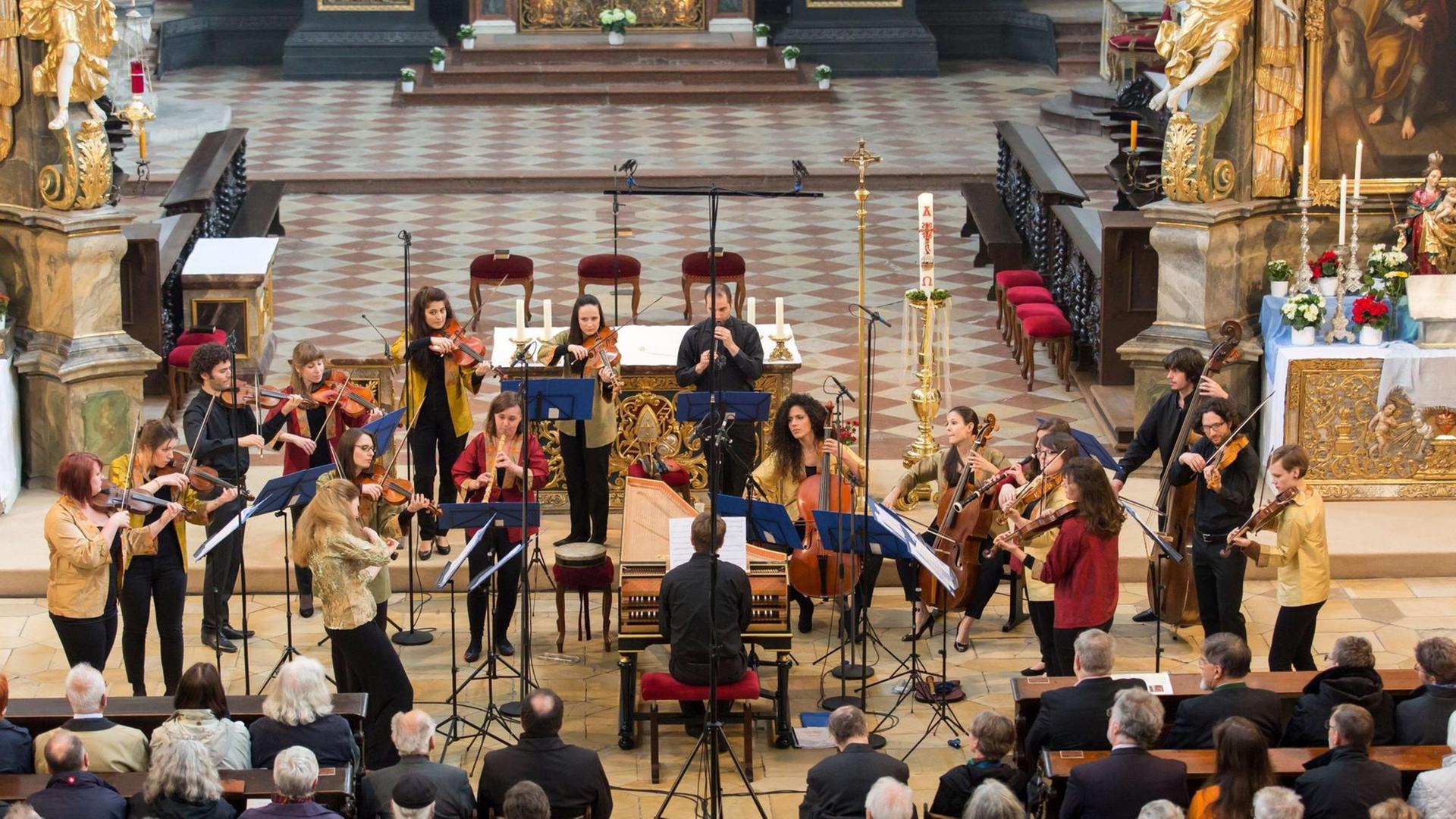 Die Musikerinnen und Musiker des Barockorchesters der Europäischen Union spielen überwiegend stehend im Altarvorraum einer Kirche. In deren Mitte sitzt der Cembalist an seinem Instrument. Der Boden ist hell gefließt, Mikrofone sind aufgestellt. Das Publikum sitzt davor auf Kirchenbänken. Die Musikerinnen und Musiker des Barockorchesters der Europäischen Union spielen überwiegend stehend im Altarvorraum einer Kirche. In deren Mitte sitzt der Cembalist an seinem Instrument. Der Boden ist hell gefließt, Mikrofone sind aufgestellt. Das Publikum sitzt davor auf Kirchenbänken.