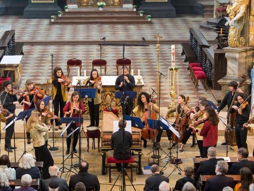 Die Musikerinnen und Musiker des Barockorchesters der Europäischen Union spielen überwiegend stehend im Altarvorraum einer Kirche. In deren Mitte sitzt der Cembalist an seinem Instrument. Der Boden ist hell gefließt, Mikrofone sind aufgestellt. Das Publikum sitzt davor auf Kirchenbänken.