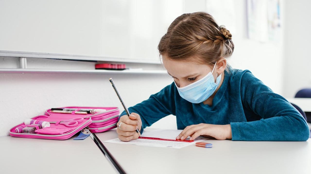 Die achtjährige Anna sitzt mit Maske an einem Tisch und bearbeitet ein Übungsblatt. Die achtjährige Anna sitzt mit Maske an einem Tisch und bearbeitet ein Übungsblatt.