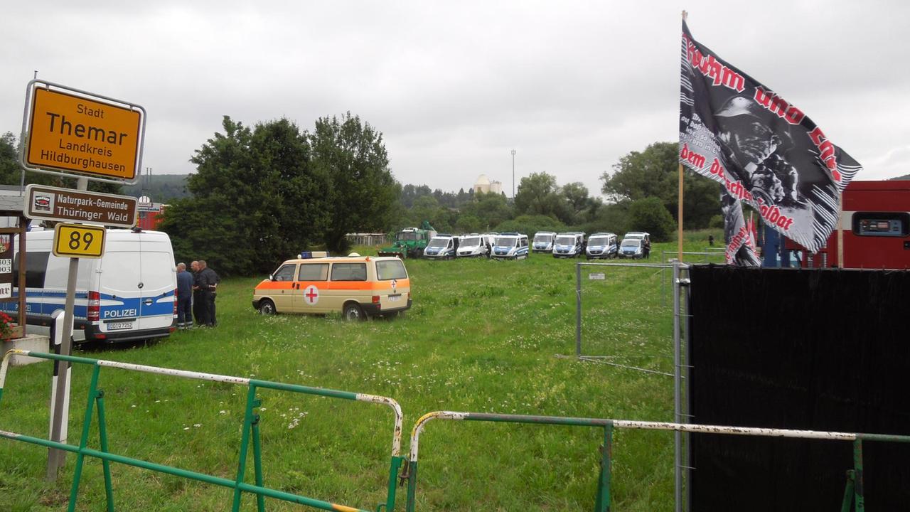 Eine Wiese mit Polizeiwagen, Absperrungen und einer Flagge mit rechtsradikalem Inhalt.
