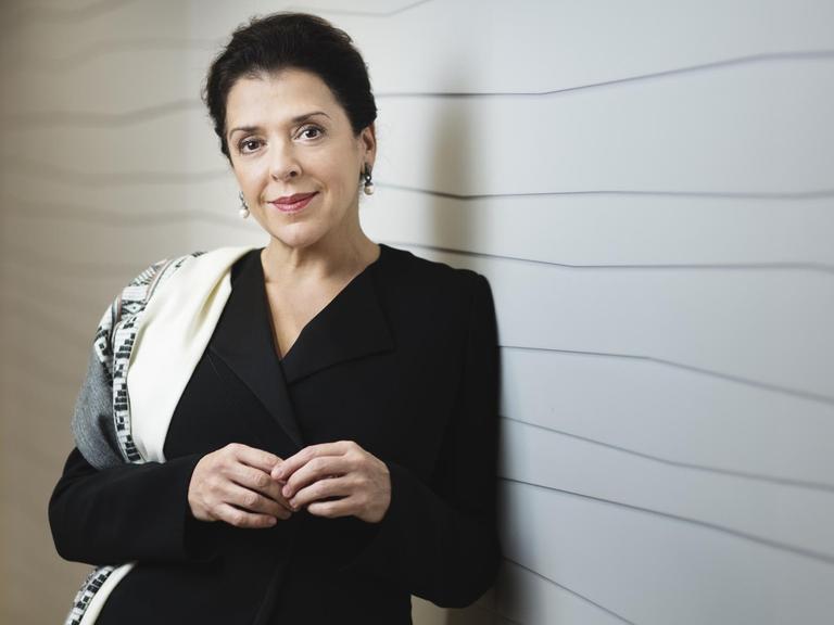 Eine Frau mit dunklen Haaren und Augen lehnt an einer weißen Wand mit schwarzen, leicht gewellten Linien, schaut in die Kamera und hält die Hände vor der dem Oberkörper leicht aneinander.