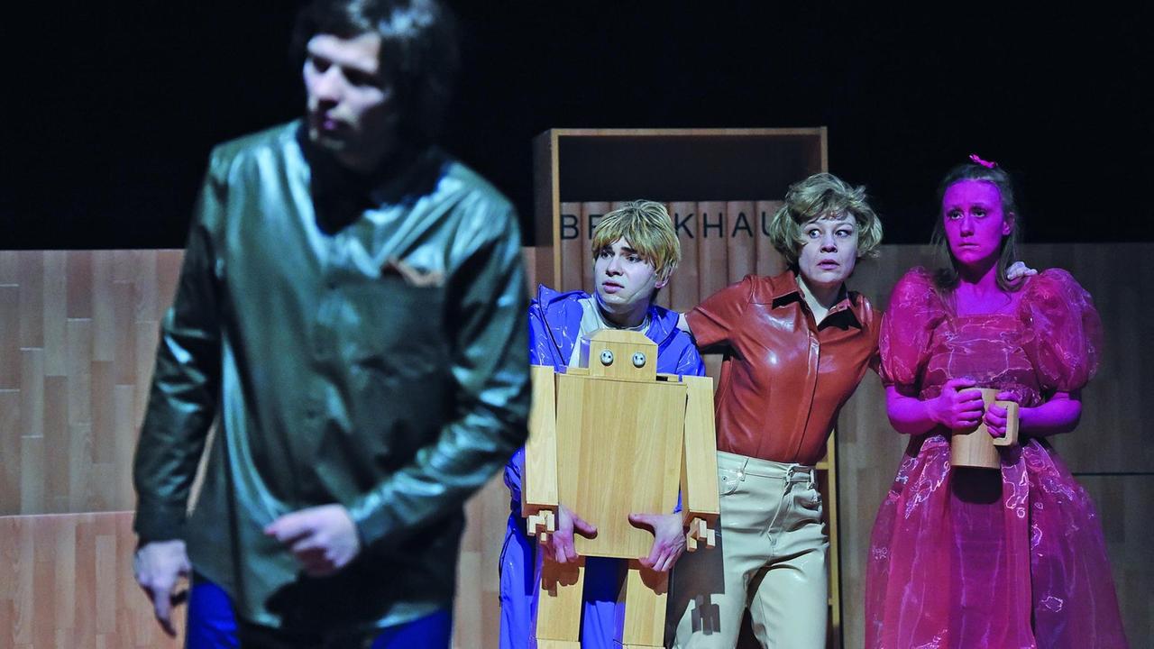 Eine Szene aus dem Theaterstück "Das Deutschland". Zu sehen sind eine Frau, die einem jungen Mann und einer jungen Frau, mit einem pinken Gesicht, an die Schulter fasst. Ein Mann im Bildvordegrung und in der Umschärfe, blickt vorsichtig zur Seite. Eine Szene aus dem Theaterstück "Das Deutschland". Zu sehen sind eine Frau, die einem jungen Mann und einer jungen Frau, mit einem pinken Gesicht, an die Schulter fasst. Ein Mann im Bildvordegrung und in der Umschärfe, blickt vorsichtig zur Seite.