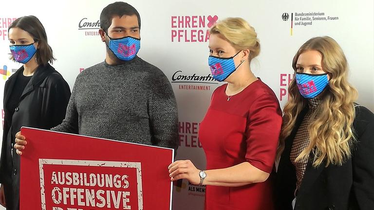 Vier Personen stehen mit Mundschutz vor einer Plakatwand. Zwei Menschen halten ein Schild, auf dem steht "Ausbildungsoffensive Pflege". Vier Personen stehen mit Mundschutz vor einer Plakatwand. Zwei Menschen halten ein Schild, auf dem steht "Ausbildungsoffensive Pflege".