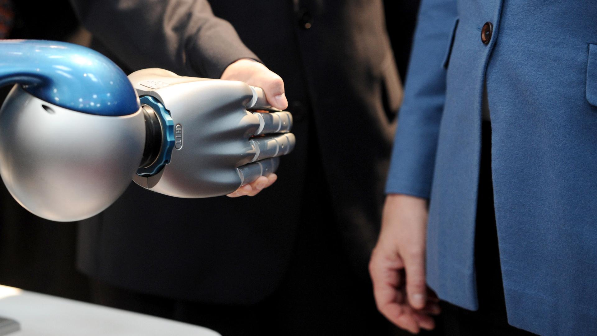 Ein Politiker schüttelt eine Roboterhand. Die Bundeskanzlerin Angela Merkel (CDU) steht daneben. Ein Politiker schüttelt eine Roboterhand. Die Bundeskanzlerin Angela Merkel (CDU) steht daneben.