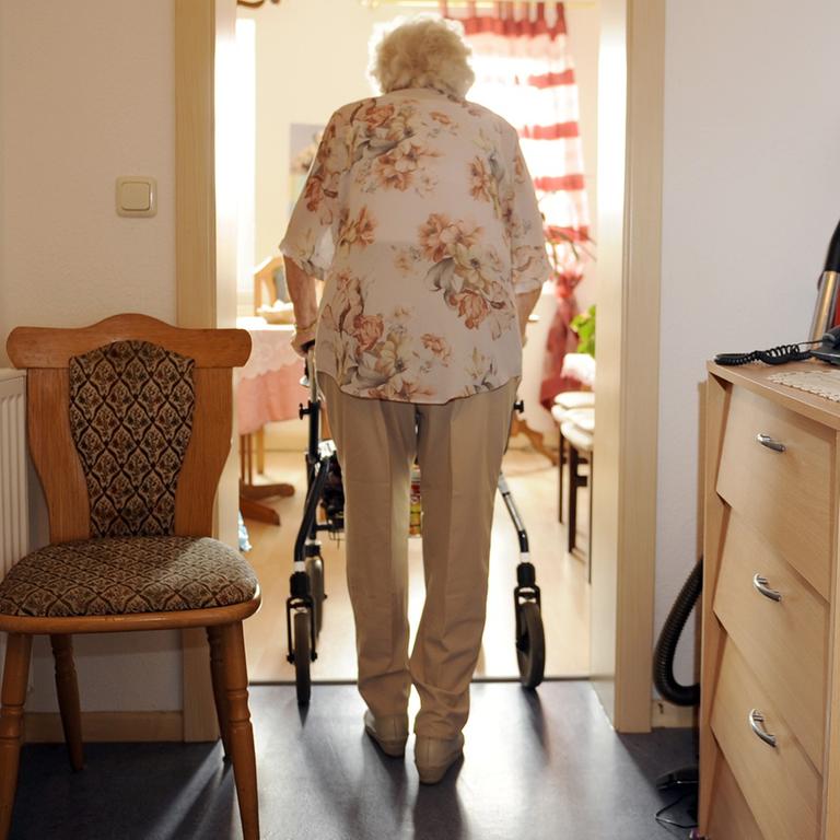 Eine ältere Frau geht am Rollator vom Wohnungsflur in ein Zimmer.