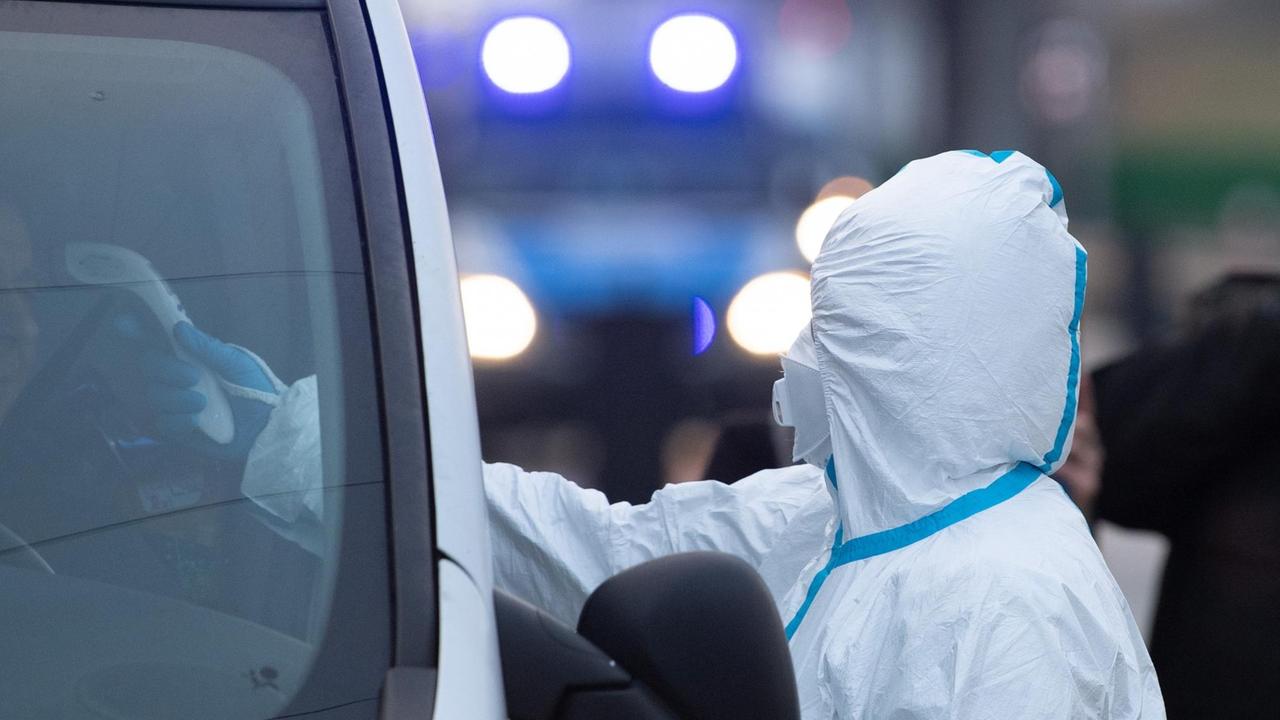 Eine Frau in weißer Schutzkleidung hält ein elektronisches Fieberthermometer durch ein geöffnetes Autofenster. Eine Frau in weißer Schutzkleidung hält ein elektronisches Fieberthermometer durch ein geöffnetes Autofenster.
