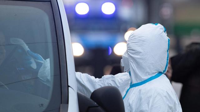 Eine Frau in weißer Schutzkleidung hält ein elektronisches Fieberthermometer durch ein geöffnetes Autofenster.