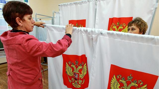 Zwei Frauen hängen in einem Wahllokal Trenn-Vorhänge mit einem Wappen zur Vorbereitung der Duma-Wahl auf.