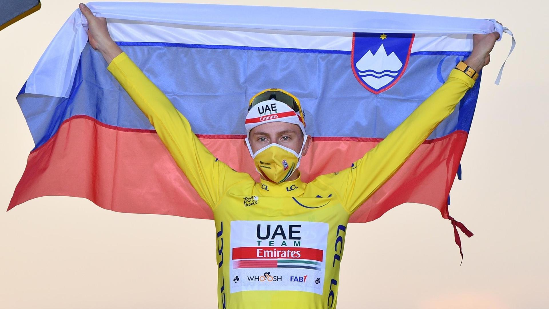 Der Renn-Fahrer Tadej Pogacar streckt im gelben Trikot die Arme nach oben und hält eine slowenische Flagge hoch.
