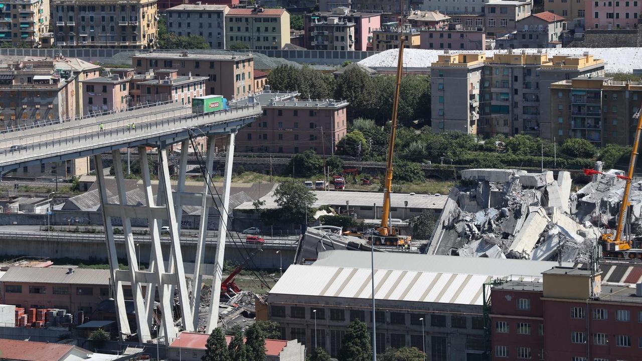 Mit Baggern und Kränen werden Trümmerteile der teilweise eingestürzten Brücke in Genua weggeräumt. Mit Baggern und Kränen werden Trümmerteile der teilweise eingestürzten Brücke in Genua weggeräumt.