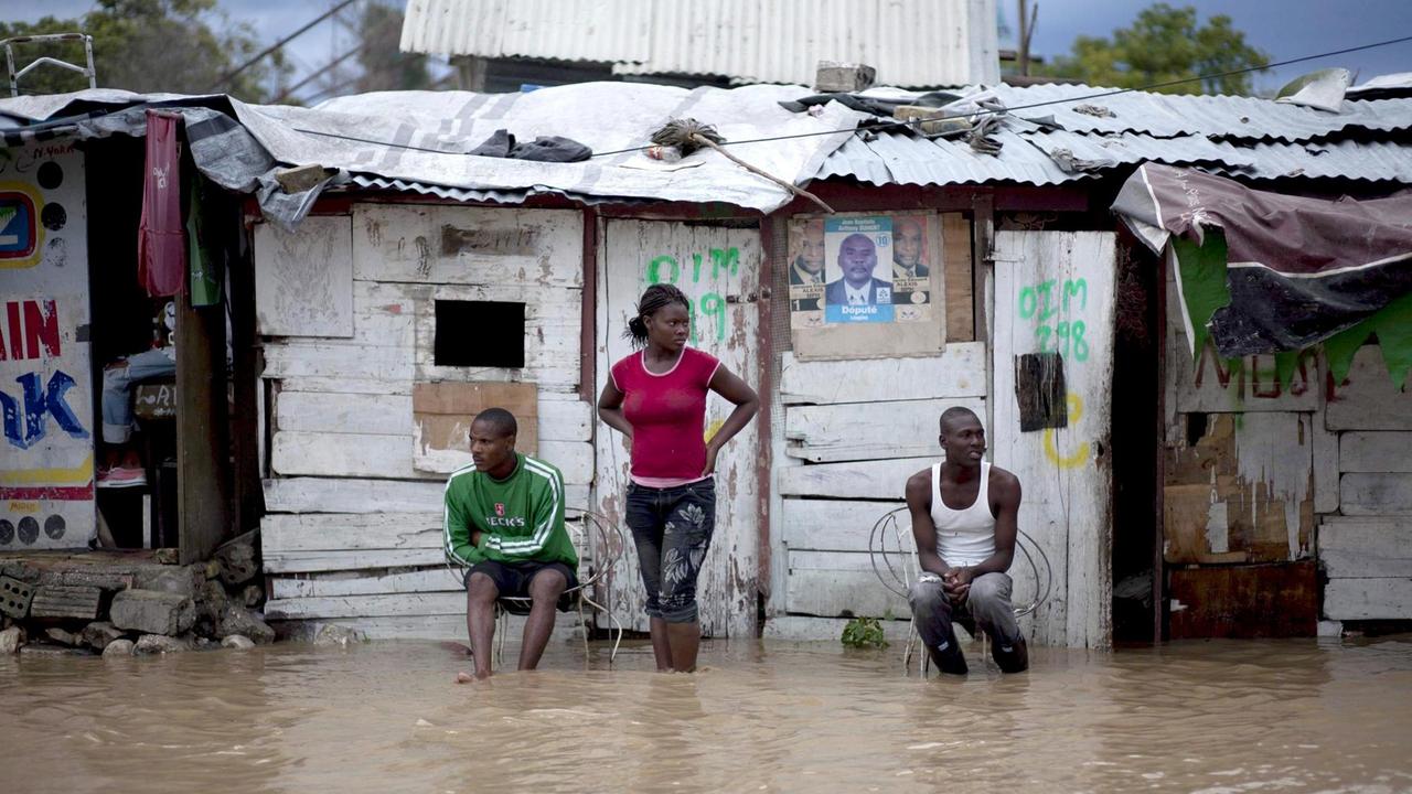 Eine Frau und zwei Männer stehen vor einer Hütte in einer überfluteten Straße in Leogane, Haiti.