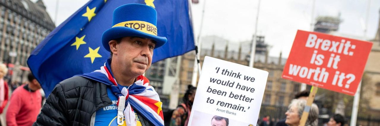 Ein Mann demonstriert mit anderen Brexit-Gegnern auf dem Londoner Parliament Square. Er trägt einen blauen Zylinder mit der Aufschrift: "Stop Brexit". Außerdem hält er ein Plakat mit einem Zitat von Arron Banks hoch: "I think we would have been better to remain." Im Hintergrund ist eine Frau zu sehen, die ein Plakat mit der Aufschrift hochhält: "Brexit: is it worth it?"
Ein Mann demonstriert mit anderen Brexit-Gegnern auf dem Londoner Parliament Square. Er trägt einen blauen Zylinder mit der Aufschrift: "Stop Brexit". Außerdem hält er ein Plakat mit einem Zitat von Arron Banks hoch: "I think we would have been better to remain." Im Hintergrund ist eine Frau zu sehen, die ein Plakat mit der Aufschrift hochhält: "Brexit: is it worth it?"