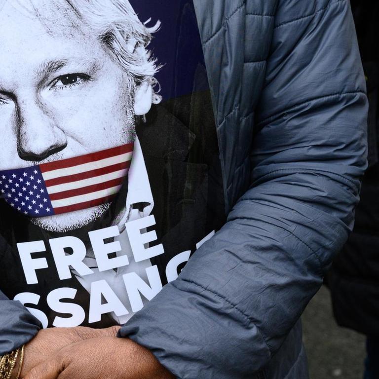 Eine Unterstützerin von Julian Assange hält vor ihrer Brust ein Schild, auf dem das Gesicht von Assange zu sehen ist. Der Mund von Assange ist auf dem Bild mit einer US-amerikanischen Flagge zugeklebt. Darunter steht auf dem Plakat "Free Assange" ("Befreit Assange").

