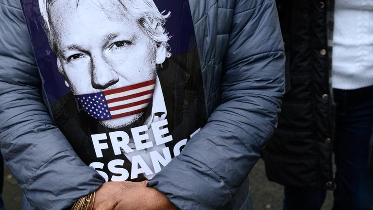 Eine Unterstützerin von Julian Assange hält vor ihrer Brust ein Schild, auf dem das Gesicht von Assange zu sehen ist. Der Mund von Assange ist auf dem Bild mit einer US-amerikanischen Flagge zugeklebt. Darunter steht auf dem Plakat "Free Assange" ("Befreit Assange").

