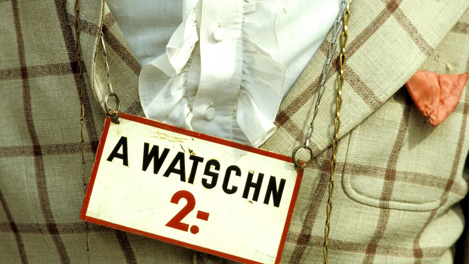 Ein Mann trägt ein Hemd mit Sakko, um den Hals hängt ihm ein Schild, auf dem steht: "A Watschn 2.-". Watschn ist ein österreichisches Wort für Ohrfeige.
