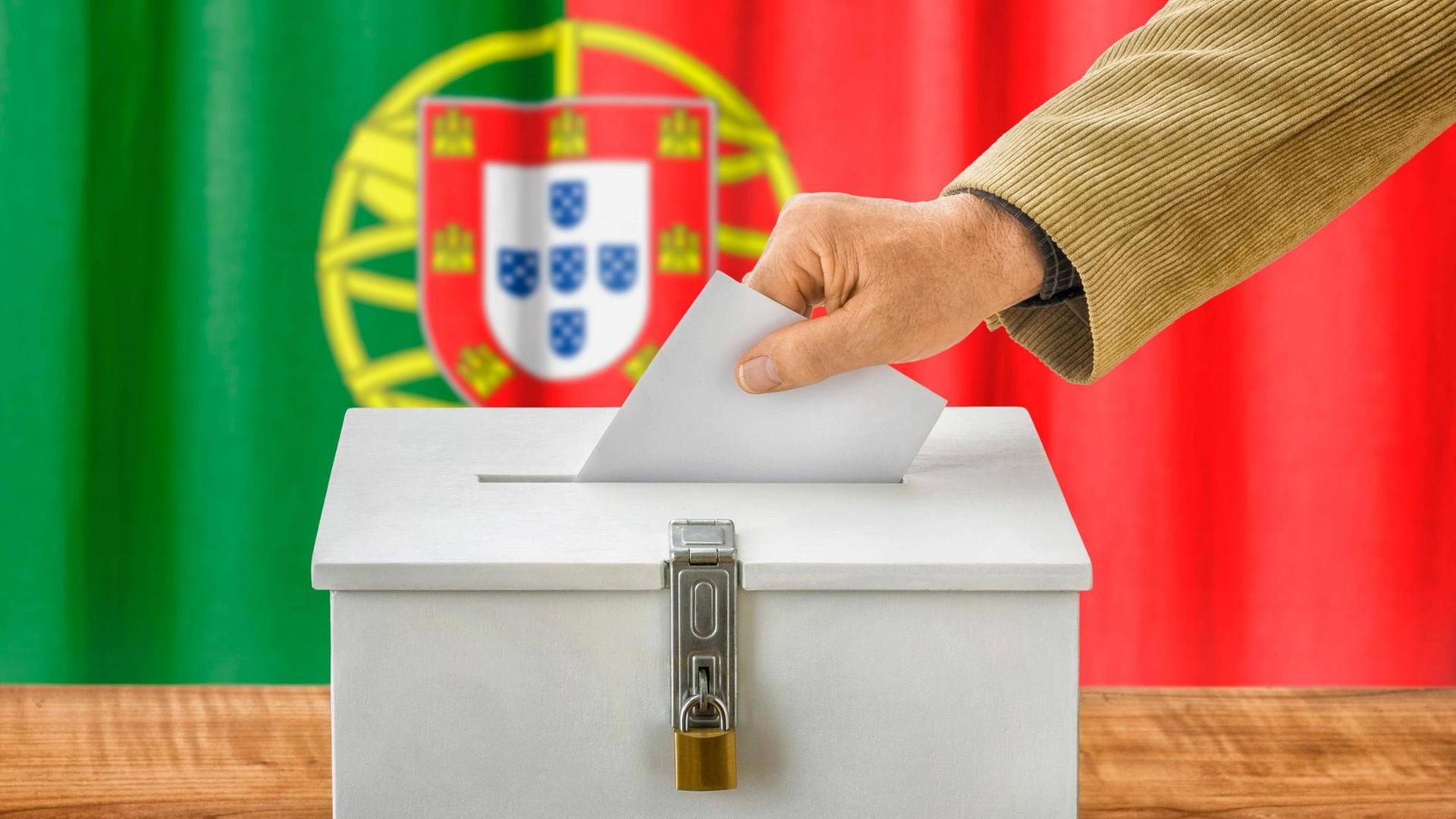 Ein Stimmzettel wird in eine Wahlurne in Portugal gesteckt, im Hintergrund ist die portugiesische Flagge zu sehen