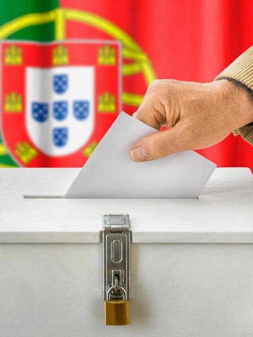 Ein Stimmzettel wird in eine Wahlurne in Portugal gesteckt, im Hintergrund ist die portugiesische Flagge zu sehen