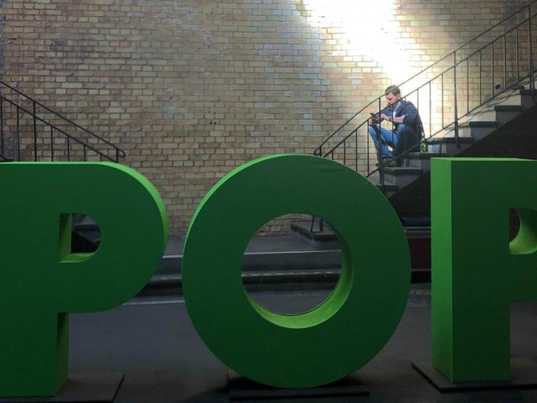 Mann sitzt auf Treppe und wird von einem Lichtschein erhellt. Im Vordergrund steht in großen grünen Buchstaben das Thema der aktuellen re:publica: “Pop”
