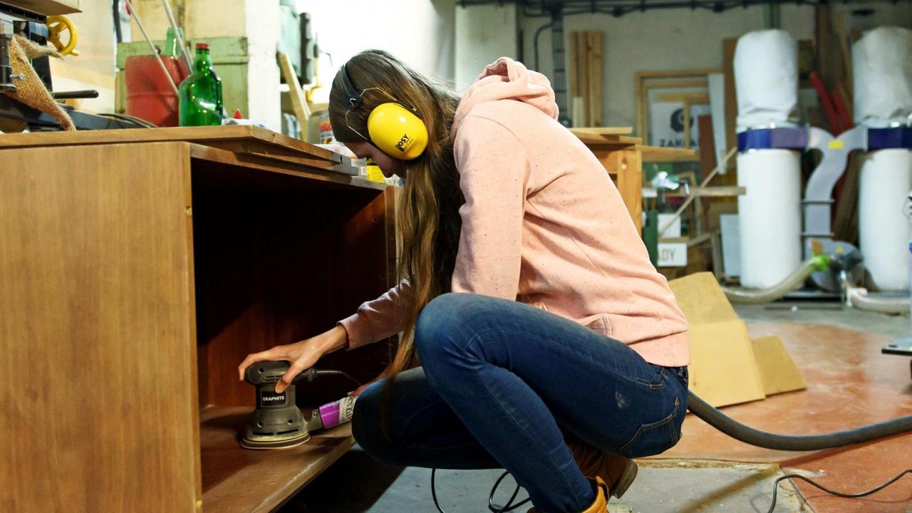 Eine Frau hockt in einer Werkstatt vor einem Sideboard aus Holz und schleift dieses mit einem elekronischen Handschleifgerät ab Eine Frau hockt in einer Werkstatt vor einem Sideboard aus Holz und schleift dieses mit einem elekronischen Handschleifgerät ab