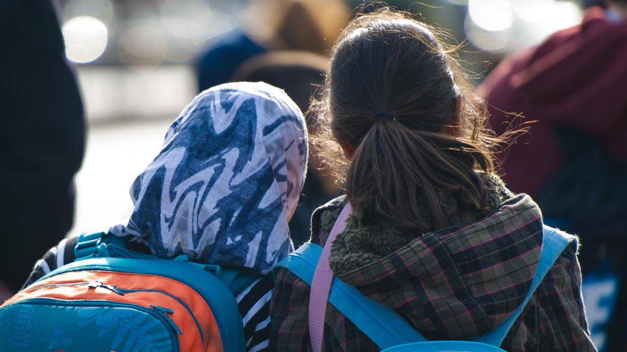 Ein Mädchen mit Kopftuch und Schulranzen steht in Berlin neben einem Mädchen ohne Kopfbeckedung.
