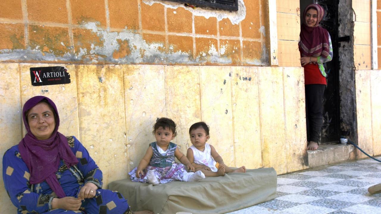 Eine Frau und zwei Kinder sitzen vor ihrem Haus in Aleppo. In der Hauswand sieht man viele Einschusslöcher. 