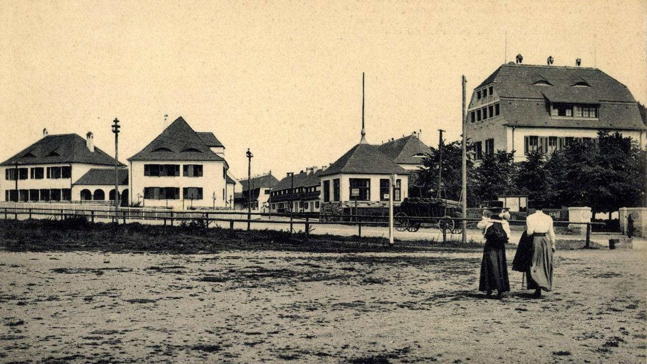 Eine historische Ansicht der Gartenstadt Hellerau. Das Schwarz-Weiß-Bild zeigt im Vordergrund zwei Damen vor den Häusern der Gartenstadt.