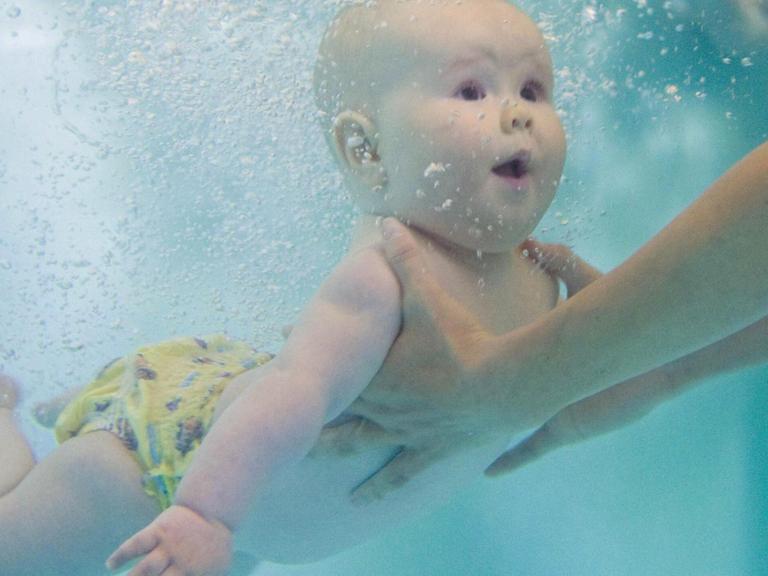 Die Unterwasseraufnahme eines Babys, das beim Schwimmunterricht unter Wasser festgehalten wird