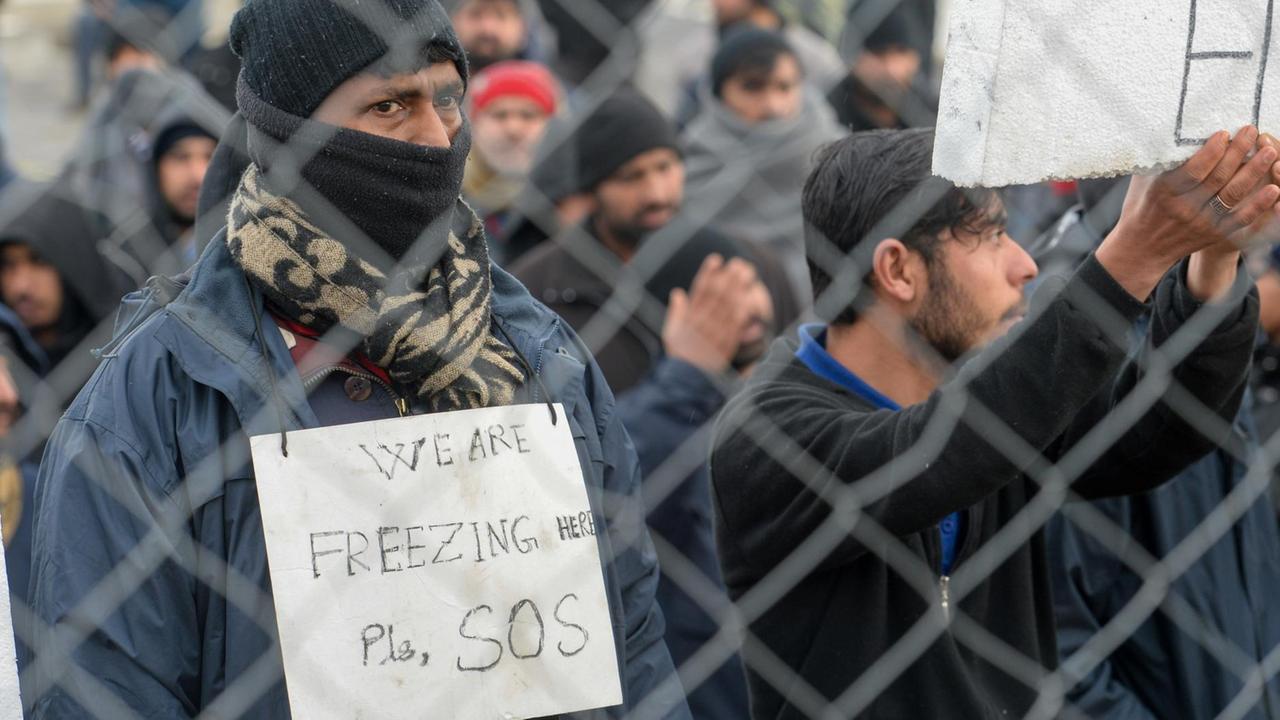 Ein Flüchling steht an einem Zaun mit einem Schild, auf dem steht: "Wir erfrieren. Rettet uns." Ein Flüchling steht an einem Zaun mit einem Schild, auf dem steht: "Wir erfrieren. Rettet uns."
